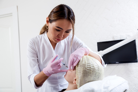 Dark-haired woman an asian beautician pushes a syringe to inject into the cheekbones of a young woman to correct the form in the medical officeの写真素材