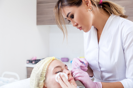 Close-up Young woman doctor cosmetician in white lab coat and sterile gloves makes injection of hyaric acid using a syringe into the cheekbones of a young woman in a medical officeの写真素材