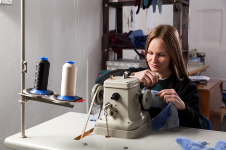 A young cheerful woman furrier smiles and sews a bright fur coat of natural blue fur on a sewing machine in the workshopの写真素材