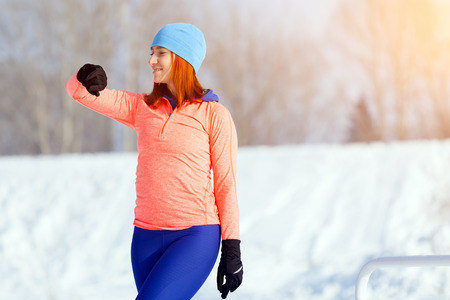 A young woman in a bright blue hat, orange sweater and elk smiles, prepares for training and looks at the sports watch before running on the playground on a bright winter dayの写真素材