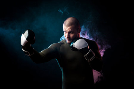 A strong dark-haired sportman  in a green sports shirt and black and white boxing gloves boxing against a blue and red vape smoke background on a black isolated の写真素材