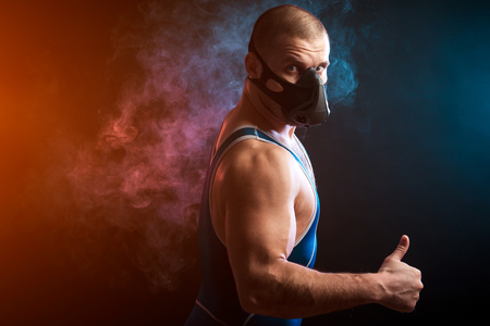 A strong dark-haired sportman  in a blue wrestling tights and training mask standing and showing thumb up  against a blue and red vape smoke background on a black isolated の写真素材