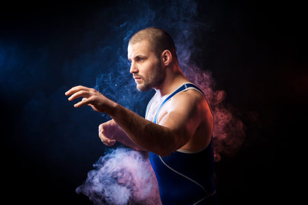 A young sporty man wrestler in a green sports shirt and blue wrestling tights  posing against a blue and red vape smoke background on a black isolated の写真素材