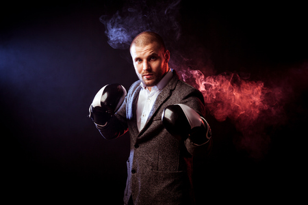 Young intelligent male businessman in white shirt and gray suit is standing in black and white boxing gloves on a background of red and blue smoke from a vape on a black isolated backgroundの写真素材