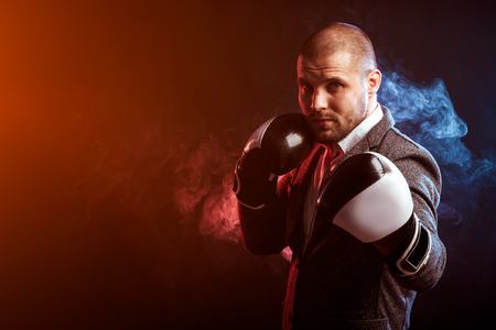 Young intelligent man businessman in white shirt, gray suit, black and white boxing gloves box in against a background of red and blue smoke from a vape on a black isolated backgroundの写真素材