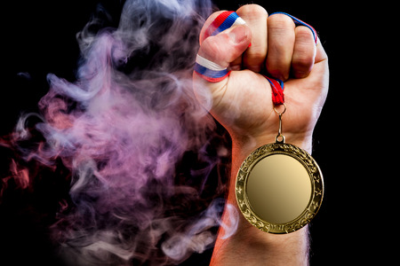 Close-up of a strong male hand holding a gold medal for a sporting achievement against a background of colored pink smoke and black isolated background. Mocap medalsの写真素材