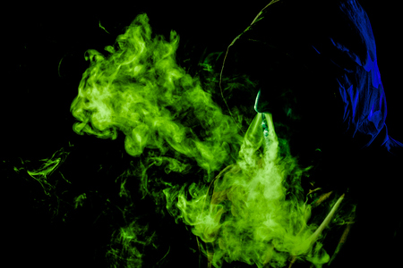 A young man in a black hood smokes a vape and exhales a colored  green and blue smoke on a black isolated backgroundの写真素材