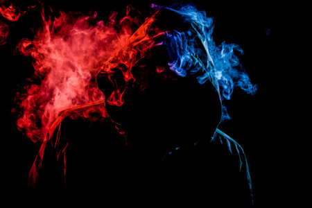 A young man in a black hood smokes a vape and exhales a colored  red and blue smoke on a black isolated backgroundの写真素材