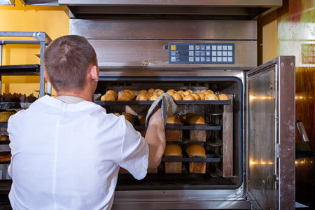 A male baker in white uniform and a beige apron bakes bread and takes cheese buns from an industrial...