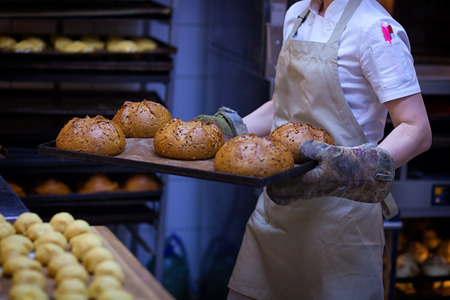 A male baker in white uniform and a beige apron takes bread from an industrial oven. Work at the bakeryの写真素材