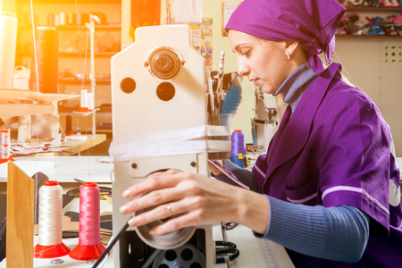 A young woman working as a seamstress in a purple unifrome sews genuine leather children's shoes on a sewing machine on a table in a sewing workshopの写真素材