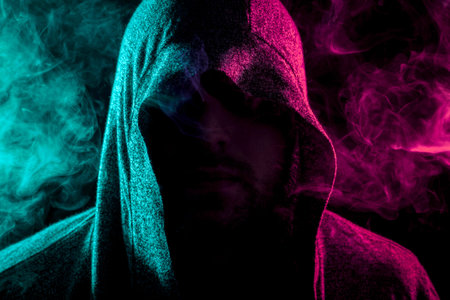 A young man in a black cape smokes and exhales large clouds of colored  green and pink smoke on a...