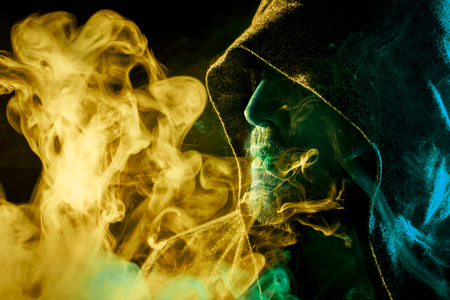 A young man in a black cape smokes and exhales large clouds of colored  yellow and blue smoke on a black isolated backgroundの写真素材