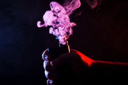 Close-up of a man holding a vape in his hand, which is smoking. Colorful smoke from cigarettesの写真素材