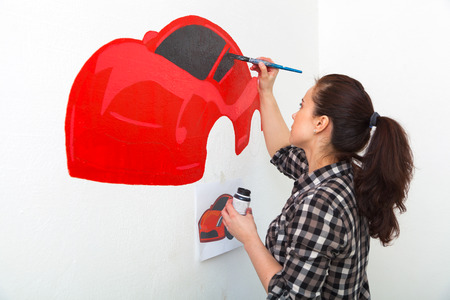 Young woman painter and mother boy draws for a child on a white wall a beautiful red car in a light children's roomの写真素材