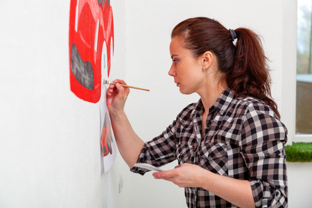 close-up of a young woman artist and mother boy drawing for a child on a white wall a beautiful red car in a light children's roomの写真素材