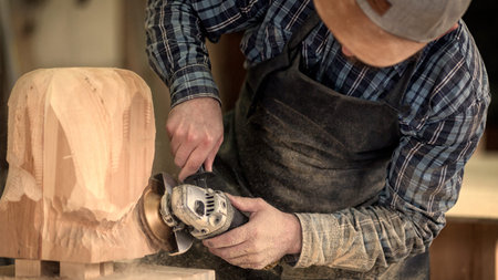 Experienced carpenter in work clothes and small buiness owner working in woodwork workshop, processes the board with an angle grinder on the table is a hammer and many tools の写真素材