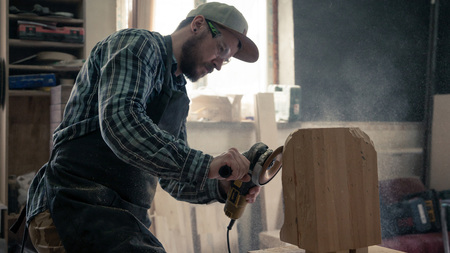Ð¡arpenter in work clothes and small buiness owner working in woodwork workshop, processes the board with an angle grinder , on the table is a hammer and many tools の写真素材
