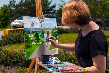 Arrtist  young woman paints on a canvas an urban landscape of a summer  with atractions of oil paints in city parkの写真素材