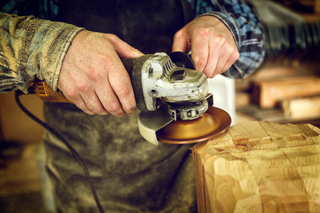 Close up experienced carpenter in work clothes and small buiness owner processes the board with an angle grinder on the table is a hammer and many toolsの写真素材
