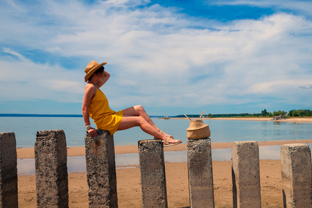 Young beautiful woman in a light casual dress and a straw hat resting and posing on rocks on a sandy beach near a blue sky on a summer day, beautiful sea against a blue skyの写真素材