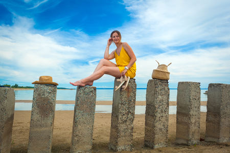 Young beautiful woman in a light casual dress and a straw hat resting and posing on rocks on a sandy beach near a blue sky on a summer day, beautiful sea against a blue skyの写真素材