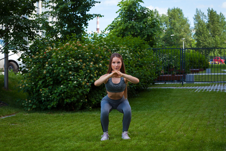 A dark-haired woman coach in a sporty short top and gym leggings shows the correct squatting technique, the position of a full squat, hands in front, on a summer day in a park on a green lawnの写真素材