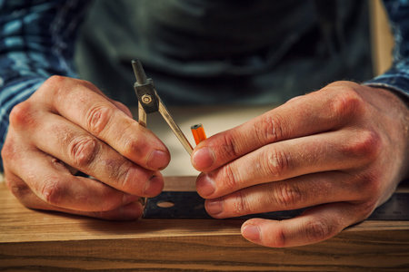 A close up of a male carpenter marks of a metal compass and a pencil on a wooden bar for making...