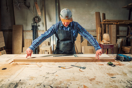 experienced carpenter in work clothes and small buiness owner measures a wooden board with a ruler and marks with pencil the necessary points for slicesの写真素材