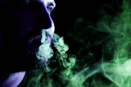 A young man exhales a cloud of colored smoke of blue and green color on a black isolated. Harm to smoking on the example of a young muggerの写真素材