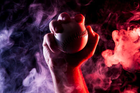 Close-up of a strong male hand holding a white baseball ball  against a background of colored  blue  and red smoke and black isolated background.の写真素材