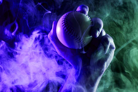 Close-up of a strong male hand holding a white baseball ball  against a background of colored  blue  and green smoke and black isolated background.の写真素材