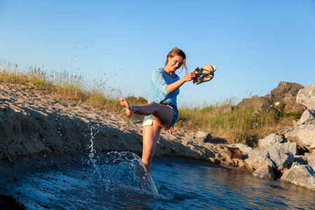 A cheerful dark-haired woman smiles, walks along the beach, kicks waves, sprays around a lot and enjoys the bright sun on a summer day. Concept of summer holidays at sea and live styleの写真素材