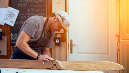 Experienced carpenter in work clothes and small buiness owner working in woodwork workshop,  using sandpaper for polishing children's helicopter blades at worktable in workshopの写真素材