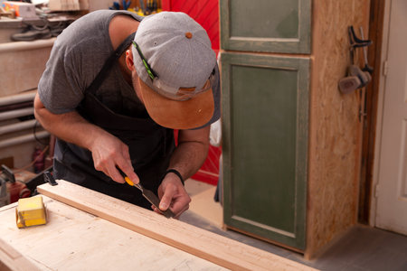 A young brunette man builder wearing green protective goggles, a gray T-shirt and a beard treating a wooden product with a chisel in the workshop, close-upの写真素材