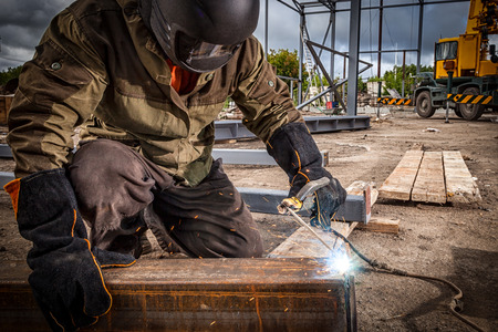 Close up of a strong man is a welder in brown uniform, welding mask and welders leathers, a metal product is welded with a arc welding machine at the construction site, blue sparks fly to the sidesの写真素材