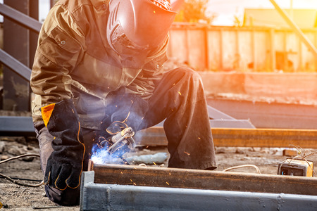 A young  man welder in brown uniform, welding mask and welders leathers, weld  metal  with a arc welding machine at the construction site, blue sparks fly to the sidesの写真素材