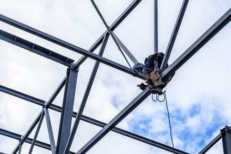 A working man is an engineer and welder in a construction overall, a welding mask is cooking metal and is sitting on a metal structure at an altitude against the blue skyの写真素材