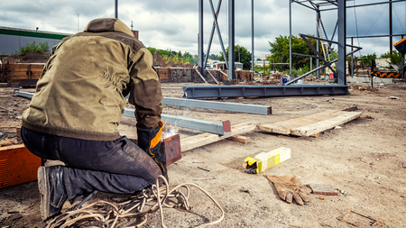 Man welder in welding mask, building uniform and blue protective gloves brews metal welding machine on street construction, in the background construction siteの写真素材