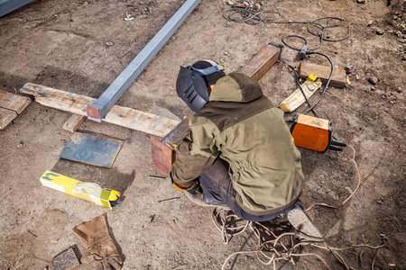 Man welder in welding mask, building uniform and blue protective gloves brews metal welding machine...