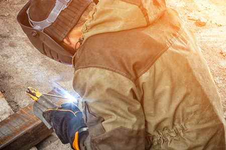 A young  man welder in brown uniform, welding mask and welders leathers, weld  metal  with a arc...