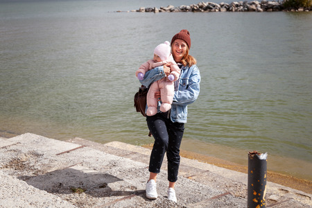 Atmospheric lifestyle photo of young beautiful  darkhaired woman  in knitting hat, in a leather jacket  and black trousers walks and plays with his little daughter  baby  in the background sea  in sunny autumn dayの写真素材