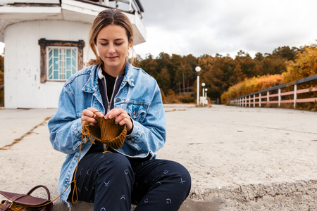 Close up of a young  married woman dressed in trendy denim coat knitting a green hat with knitting needles  against the background of the old lighthouse  in sunny autumn day . Freelance creative working and living conceptの写真素材