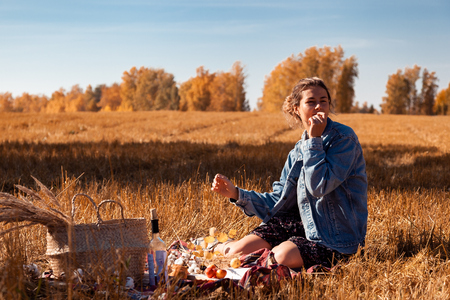 Picnic in the fresh air: a young woman in a jeans jacket and a dress eating an apple and enjoying nature, sitting on a plaid with a picnic basket, apples, wine. On the back of the background is the autumn landscape of the field.の写真素材