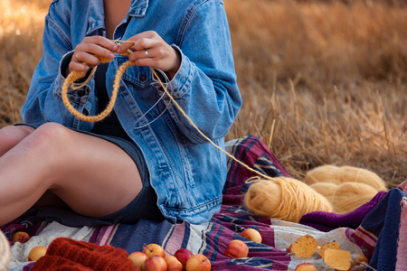 Concept of a freelancer work in the open air.  A young woman in a denim jacket and dress knitting hat with needle on plaid with a picnic basket, apples, wine on  the  background autumn  field.の写真素材