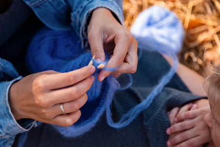 Concept of a freelancer work in the open air. Close up  of a young woman in a denim jacket knitting blue hat with needle on plaid.の写真素材