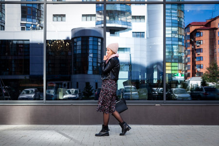 Portrait of a beautiful young model in blue knitted hat standing do to work, on background mirrow wall on  sunny autumn day in  big city. Autumn warm photo.Woman smiling and look away, joyful cheerful mood.の写真素材