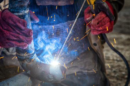 Man welder in welding mask, building uniform and red protective gloves brews metal welding machine...