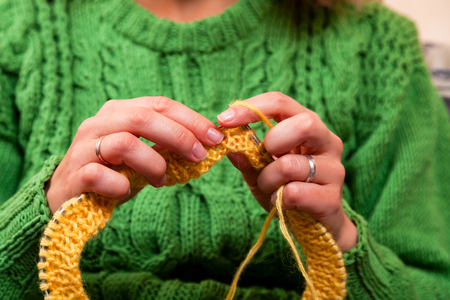 Close-up of a beautiful woman in knitting green sweater knitting needles from a natural woolen thread a yellow hat, a woman shows how to knit correctlyの写真素材