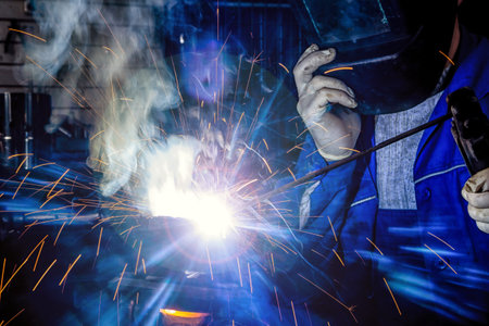 Man welder in welding mask, building uniform and protective gloves welds metal car part with welding...